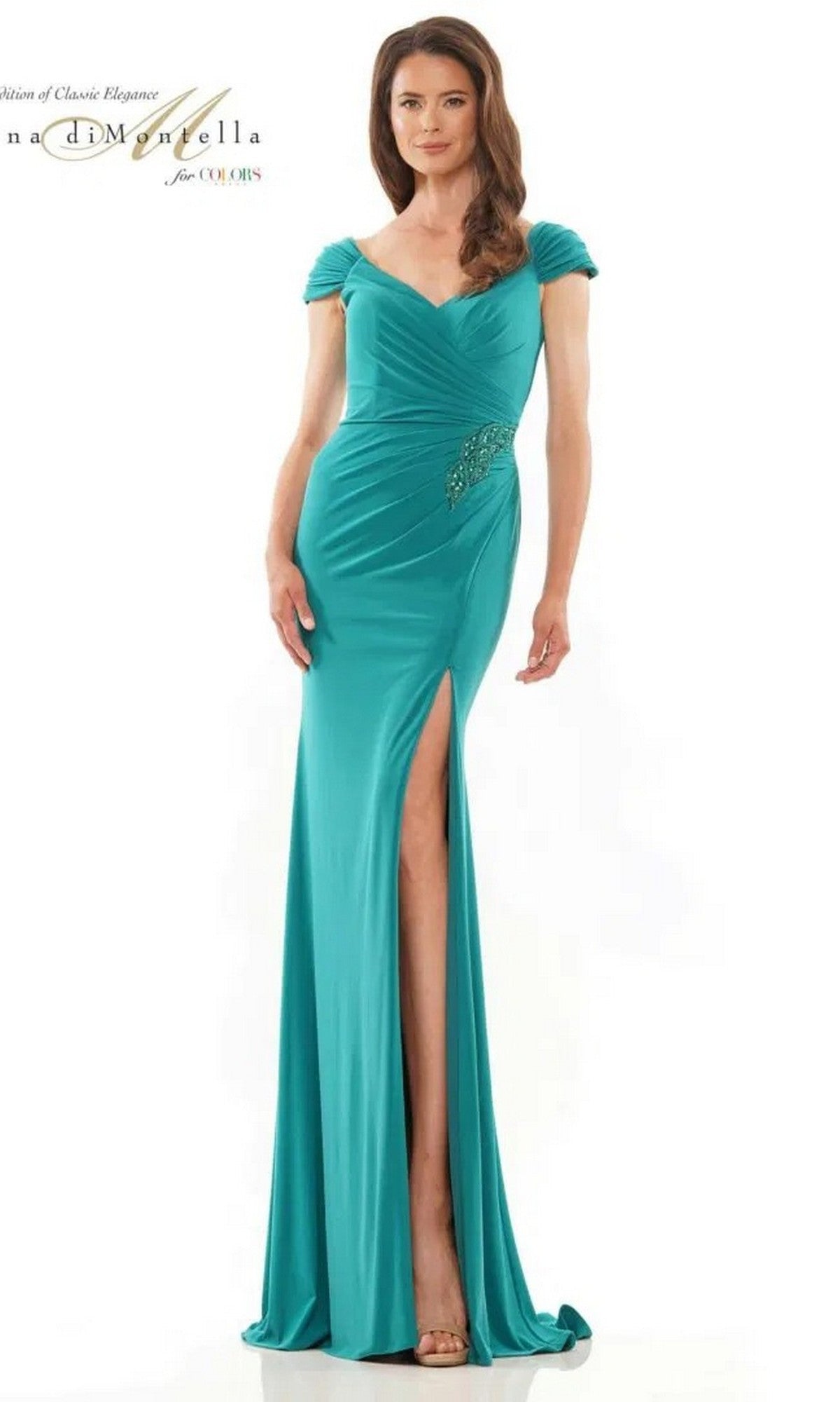 A woman in an emerald Long Formal Dress: Rina Di Montella RD2824, featuring a floor-length jersey design with a high-leg slit and short sleeves, stands against a plain white background.
